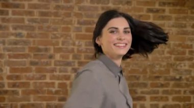 Genç güzel kadın kamera seyir ve çevresinde büküm, atlama ve gülüyor, mutlu, kısa saçlı kahverengi tuğla duvar arka plan