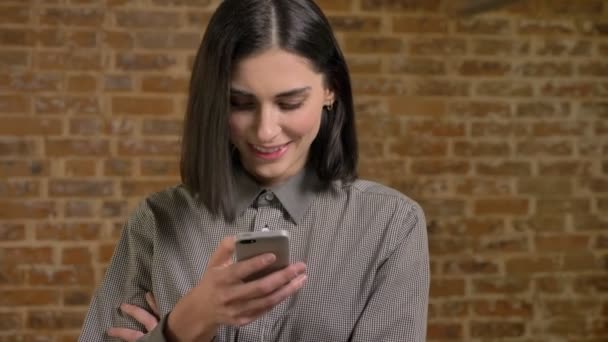 Portrét mladé krásné ženy s krátké hnědé vlasy textové zprávy na telefonu a usmíval se, šťastná, cihlová zeď na pozadí — Stock video