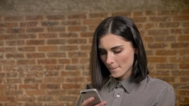 Retrato de la joven mujer bonita mensajes de texto en el teléfono y morder los labios, concentrado, fondo de la pared de ladrillo — Vídeo de stock