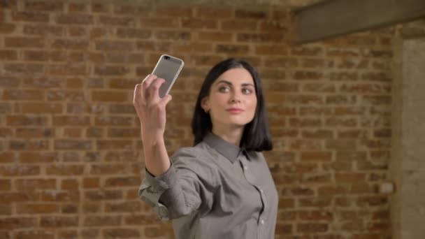 Ung vacker kvinna med kort brunt hår tar selfie med telefon och poserar för kameran, tegel vägg bakgrund — Stockvideo