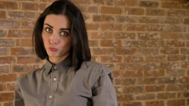 Jeune femme charmante aux cheveux bruns courts posant pour caméra et faisant des visages drôles, fond de mur de briques — Video