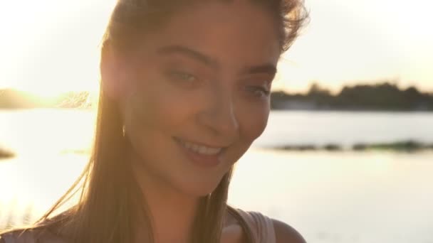 Πορτρέτο του όμορφη γυναίκα ψάχνει στην κάμερα και να χαμογελά, ηλιοβασίλεμα και φακού φωτοβολίδα, Ποταμός και η φύση φόντο — Αρχείο Βίντεο