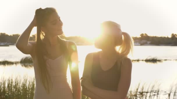 Dvě mladé krásné dívky stojící na pláži při pohledu na sebe a pak za zavřenými, s úsměvem, objektiv odlesku, řeka a přírodní pozadí