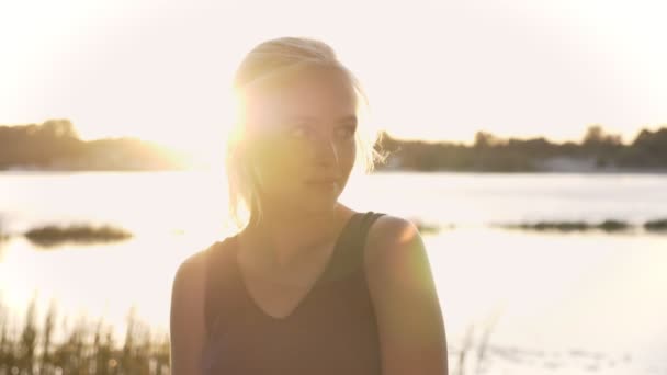 Retrato de una joven rubia mirando a cámara, tocando el cabello y sonriendo, puesta de sol y destello de lente, fondo de río y naturaleza — Vídeo de stock