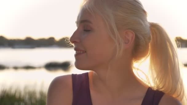 Retrato de jovem loira olhando para a câmera, sorriso bonito, sol brilhando brilhantemente, rio e natureza fundo — Vídeo de Stock