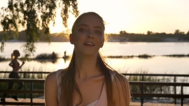 Portret van jonge vrouw op zoek zijwaarts en glimlachend, andere vrouw draait op rug, lens flare, rivier en natuur achtergrond — Stockvideo