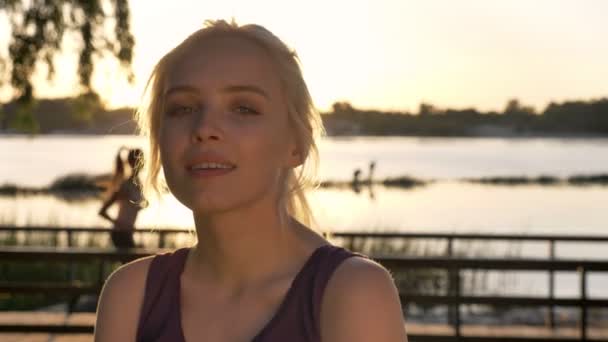 Porträt einer jungen blonden Frau, die in die Kamera blickt und lächelt, Frau läuft auf dem Rücken, Sonnenuntergang, Fluss und Natur Hintergrund — Stockvideo