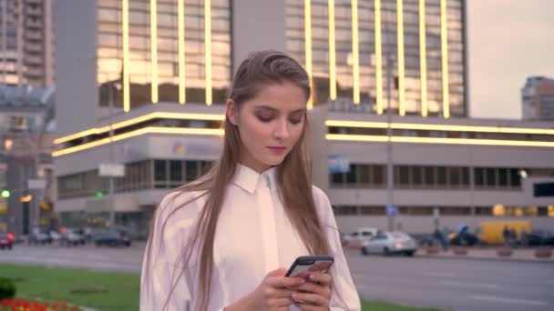 Giovane bella ragazza seria sta digitando il messaggio sul suo smartphone al tramonto nel centro della città in estate, processo di pensiero, concetto di comunicazione — Video Stock