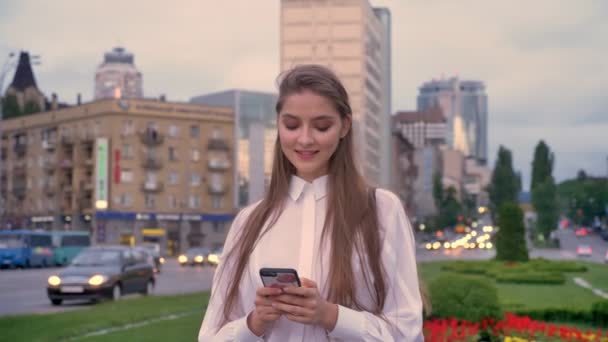 Молода щаслива дівчина друкує повідомлення на своєму смартфоні на заході сонця в центрі міста влітку, думаючи концепцію, концепцію спілкування — стокове відео