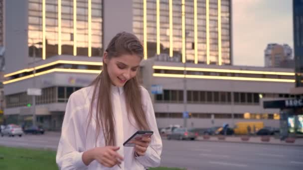 Молода красива дівчина друкує повідомлення на своєму смартфоні на заході сонця в центрі міста влітку, думаючи концепцію, концепцію спілкування — стокове відео