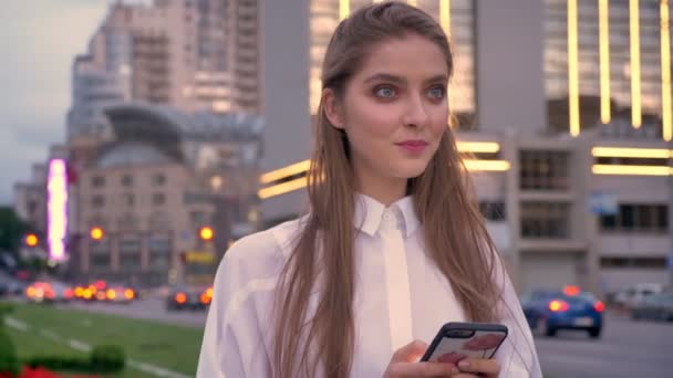 Giovane ragazza carina sta guardando sul suo smartphone al tramonto nel centro della città in estate, concetto di pensiero, concetto di comunicazione — Video Stock