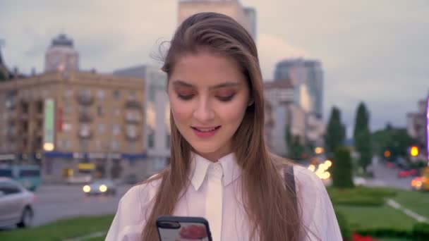 Joven hermosa chica concentrada está escribiendo mensaje en su teléfono inteligente al atardecer en el centro de la ciudad en verano, sonriendo, concepto de comunicación — Vídeos de Stock