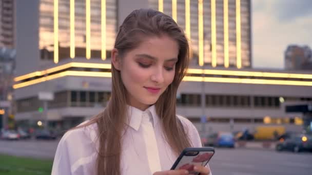 Aantrekkelijke geconcentreerde meisje is bericht in te typen op haar smartphone bij zonsondergang centrum in de zomer, glimlachen, communicatieconcept — Stockvideo