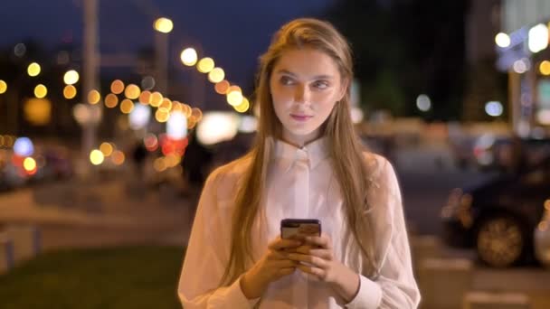 Joven chica atractiva está escribiendo mensaje en su teléfono inteligente en la noche en el centro de la ciudad en verano, concepto de pensamiento, concepto de comunicación — Vídeos de Stock