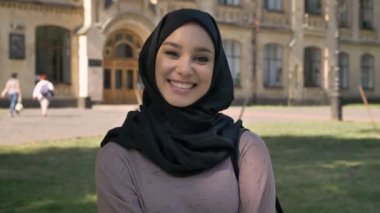 Genç Müslüman kıza hijab ayakta ve yaz aylarında gündüz gülümseyerek kameraya, Bina arka plan, religiuos kavramı, duygusal kavramı üzerinde izliyor