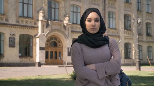 Young serious muslim girl is standing with arms crossed in daytime in summer, watching at camera, building on background, religious concept — Stock Video