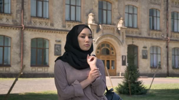 年轻严肃的穆斯林女孩在面纱是站立和固定她的围巾在白天在夏天, 观看在照相机, 修造在背景, 宗教概念 — 图库视频影像