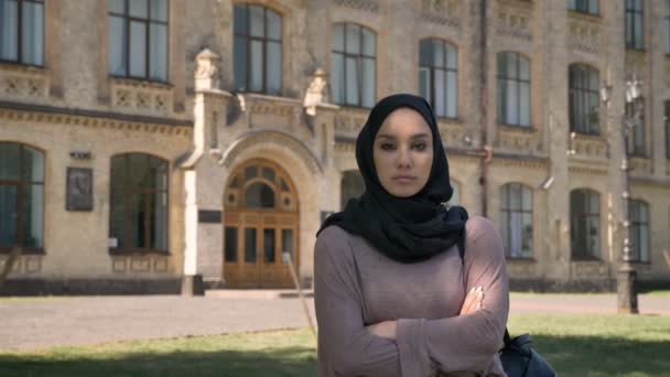 Young self-confident muslim girl is standing with arms crossed in daytime in summer, watching at camera, building on background, religious concept — Stock Video