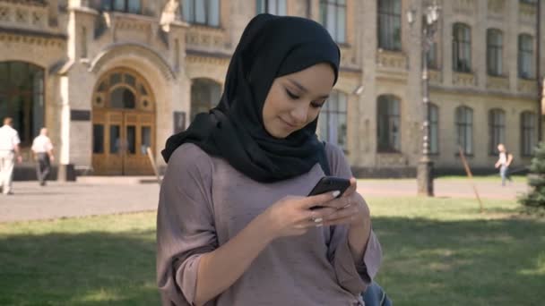 Giovane ragazza musulmana dolce in hijab sta digitando il messaggio su smartphone e sorridendo di giorno in estate, costruendo su sfondo, concetto religiuos, concetto di comunicazione — Video Stock