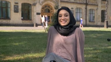 Genç Müslüman kız hijab kamera, arka plan, dini kavramı, üzerinde builging izlerken güneşin altında'nın bahçesinde oturuyor mutlu kavramı relax.