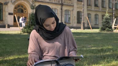 Genç Müslüman kız hijab çimlerde oturuyor ve dergisi okuma, builging arka planda, dini kavramı, rahatla kavramı