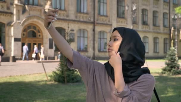 Jovem linda garota muçulmana no hijab está fazendo selfie em seu smartphone durante o dia no verão, construindo em segundo plano, conceito religiuos, conceito de comunicação — Vídeo de Stock