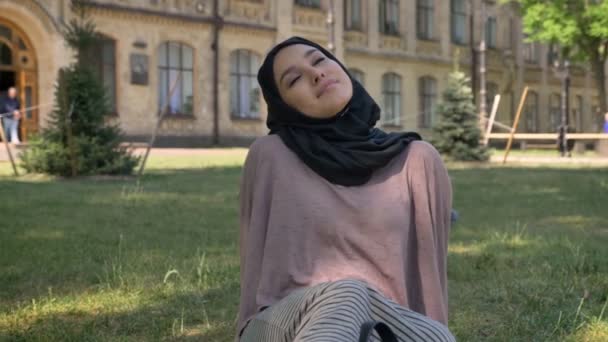 Unga vackra muslimsk flicka drömmare i hijab sitter på gräsmattan, builging på bakgrund, religiösa begrepp, koppla av koncept — Stockvideo