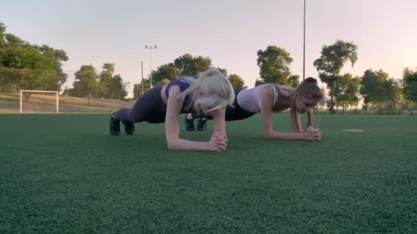 Две молодые красивые женщины делают упражнения на доске на футбольном поле в парке в течение утра, фитнес-модели упражнения — стоковое видео