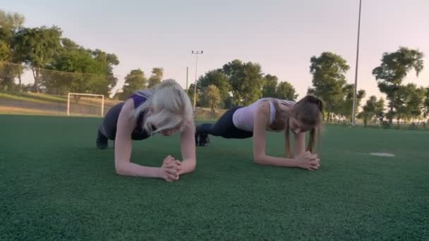 Двое молодых красивых женщин делают упражнения на доске на футбольном поле в парке утром, потеря веса — стоковое видео