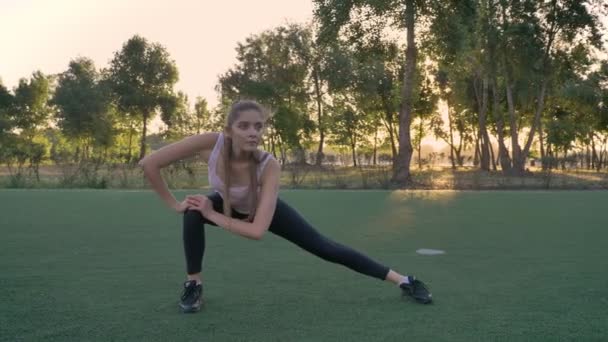 Молодая красивая женщина растягивается на футбольном поле в парке во время захода солнца, фитнес-модель упражнения — стоковое видео