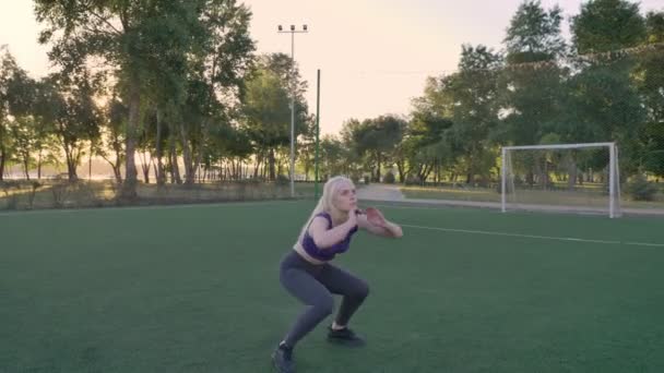 Молода блондинка робить присідання стрибає вправи на футбольному полі в парку, ранкова робота — стокове відео
