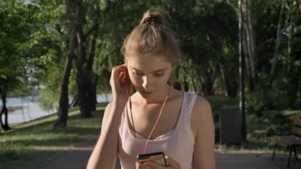 Jonge mooie vrouw dragen van oordopjes en starten lopen in park tijdens de ochtend, glimlachend — Stockvideo
