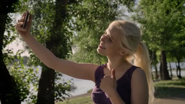 Portré, fiatal szőke nő vesz selfie neki a telefon, hüvelykujj felbukkan, és mosolyogva a kamera, a park közelében folyó háttér — Stock videók
