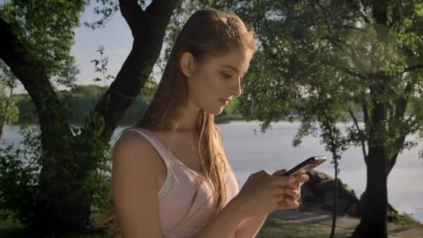 Молода красива жінка друкує на телефоні, дивлячись в камеру в парку біля річки і усміхнений, красивий вид — стокове відео