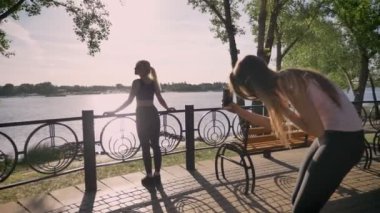 İki genç kadının çekim, onlardan biri parkta duran ve poz, diğer telefon, Nehri yakınında park arkadaş tutarken