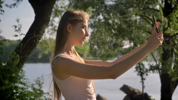 Jonge mooie vrouw die selfie met haar telefoon, kijken in de camera en lacht, permanent in park in de buurt van rivier — Stockvideo