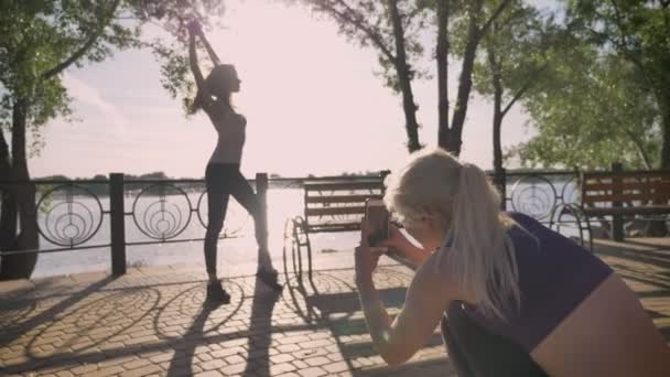 Två kvinnliga vänner i parken tar bilder, en av dem står och poserar för kameran, medan andra håller telefonen, park nära floden — Stockvideo