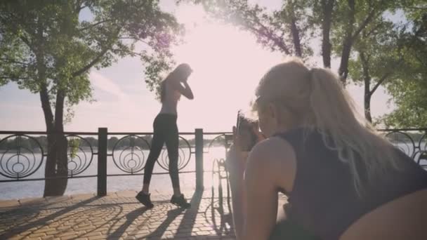 Blond kvinna tar bilder av hennes vän, andra kvinna poserar för fotot, vacker utsikt, park nära floden bakgrund — Stockvideo
