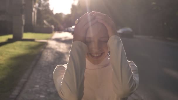 Молодая счастливая необыкновенная смелая девушка стоит на улице в дневное время летом, касаясь головы, улыбаясь, наблюдая за камерой — стоковое видео