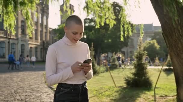 Έκτακτη τολμηρή κοπέλα είναι το περπάτημα μέσα από το πάρκο στο φως της ημέρας, το καλοκαίρι, πληκτρολογώντας το μήνυμα στο smartphone, βλέποντας στο έννοια επικοινωνίας κάμερα, κτίριο σε φόντο — Αρχείο Βίντεο