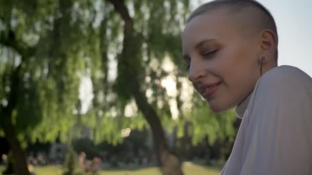 Gelukkig buitengewone meisje draait hoofd en horloges op camera in park in daglicht in de zomer, glimlacht, flirt concept, blured achtergrond — Stockvideo