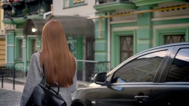 Mujer joven con el pelo largo de jengibre caminando en la calle y la celebración de la mochila en un hombro, fondo del edificio verde, vista trasera — Vídeos de Stock