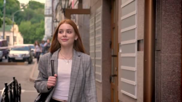 Молодая очаровательная рыжая женщина гуляет по городу и смотрит боком, улыбаясь, держа рюкзак на одном плече — стоковое видео