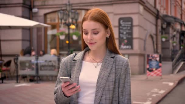 Joven bonita mujer de jengibre de pie en la calle de la ciudad y escribiendo en el teléfono, sonriendo, edificio con fondo de café — Vídeos de Stock