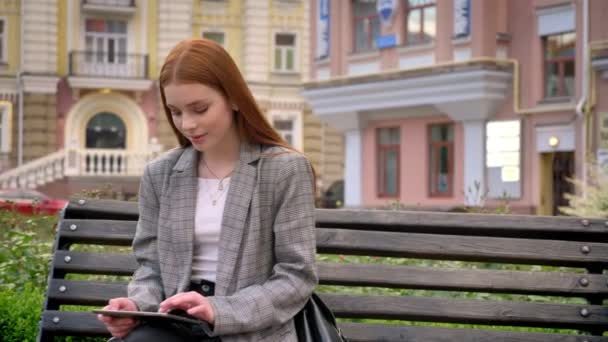 Joven bonita mujer de jengibre en chaqueta sentado en el banco y el uso de la tableta, fondo de la calle de la ciudad — Vídeos de Stock
