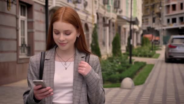 Młody imbir uroczy kobieta stoi na ulicy, patrząc na telefon i w aparacie, trzymając uprzęży plecaka, uśmiechnięty, budynek w tle — Wideo stockowe