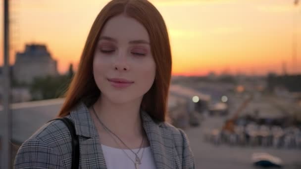 Retrato de mujer jengibre joven en chaqueta de pie en el puente y mirando a la cámara, sonriente, fondo de fábrica industrial, puesta de sol — Vídeos de Stock