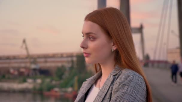 Портрет молодой рыжей женщины, стоящей на мосту, смотрящей в сторону и затем в камеру, улыбающейся, закат — стоковое видео