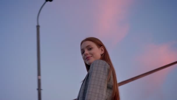 Young beautiful woman with long ginger hair looking in camera and turning around, blue sky above, circling around view, low angle — Stock Video