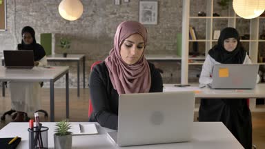 Oturma ve çalışma modern office, kamera seyir ve gülümseyen Müslüman kadın işçi hijab içinde üç genç Müslüman kadının